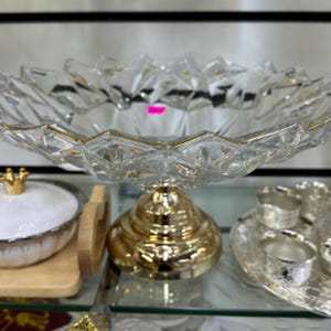 Op de afbeelding: Een heldere glazen serveerschaal met een gouden rand en een goudkleurige voet. De kom heeft een decoratief, facetgeslepen ontwerp. Andere items zijn een wit en goudkleurige kom met deksel en een zilveren theeservies.