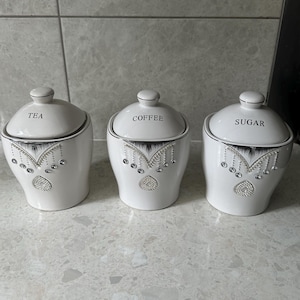 Peut inclure: Trois pots en céramique blanche avec des inscriptions noires et des accents argentés. Les pots sont étiquetés "Thé", "Café" et "Sucre".