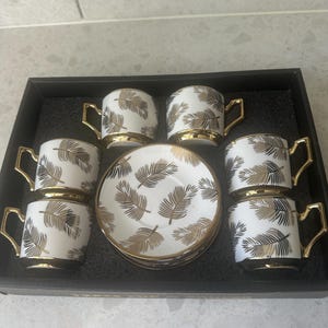 Peut inclure: Un service à café de six pièces en céramique blanche avec un motif de plumes dorées. Les tasses ont des anses et des bords dorés. L'ensemble est présenté dans un plateau noir.