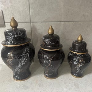 May include: Three decorative black ceramic jars with lids. The jars have a marble-like pattern with white and gold veining. Each jar has a gold-colored base and a gold finial on the lid. The jars are of varying sizes.
