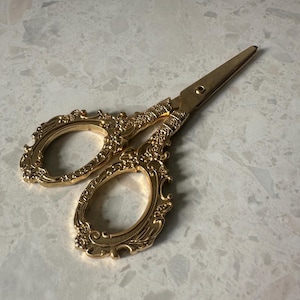 May include: An ornate pair of gold-coloured scissors. The handles and blades feature a detailed, vintage design with floral and scrollwork patterns. The blades are sharp and pointed. The scissors are open, ready for use.
