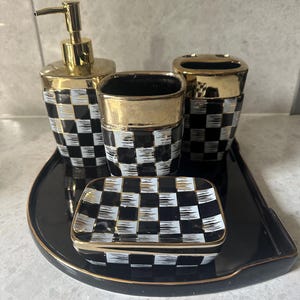 Op de afbeelding: Een badkamerset met een zwart-wit geblokt patroon en gouden accenten. De set bevat een zeepdispenser, een beker, een tandenborstelhouder, een klein schaaltje en een zwart dienblad met een gouden rand.