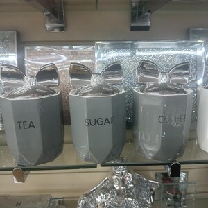 Könnte beinhalten: Drei graue, facettierte Behälter mit silbernen Schleifendeckeln. Die Behälter sind mit "TEA", "SUGAR" und "COFFEE" in schwarzem Text beschriftet. Die Behälter werden auf einem Glasregal ausgestellt.