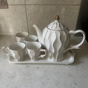 Puede incluir: Un juego de té de cerámica blanca con detalles dorados. El juego incluye una tetera, cuatro tazas y una bandeja. La tetera tiene un adorno dorado y un diseño ondulado. Las tazas y la tetera tienen ribetes dorados.