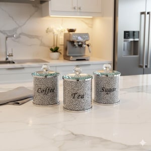 Puede incluir: Tres botes de cocina plateados con tapas de cristal, etiquetados como "Coffee", "Tea" y "Sugar". Los botes tienen un exterior brillante y están sobre una encimera blanca. La cocina tiene un diseño moderno.
