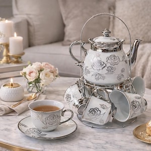 Könnte beinhalten: Weißes Porzellan-Teeservice mit silbernen Akzenten und Rosenmuster. Das Set umfasst eine Teekanne, Tassen und Untertassen, die auf einem runden Tisch mit Marmorplatte angeordnet sind. Die Szene wird durch Blumen, Kerzen und Gebäck ergänzt.