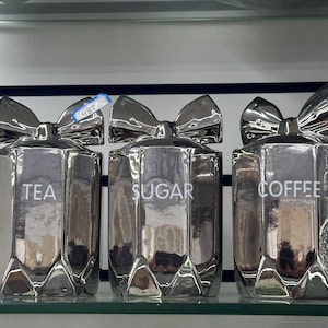 May include: Three silver canisters with a bow design and labels that read "TEA", "SUGAR", and "COFFEE".