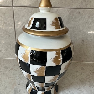 Pode incluir: Um vaso decorativo de cerâmica com tampa. O vaso é pintado com um padrão de xadrez em preto e branco, acentuado com pinceladas e acabamentos dourados. A tampa é encimada por um remate dourado.