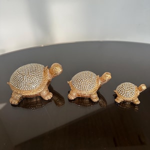 Può includere: Tre statuette decorative a forma di tartaruga color oro. Ogni tartaruga ha un guscio ricoperto di piccoli cristalli scintillanti. Le tartarughe variano di dimensioni, la più grande misura circa 15 cm di lunghezza.