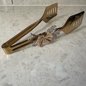 Könnte beinhalten: Goldfarbene Servierzange mit geschlitzten Enden und einem dekorativen floralen Akzent. Die Zange ist aus Metall gefertigt und zum Servieren von Speisen bestimmt. Der florale Akzent ist eine Mischung aus Gold- und Silbertönen.