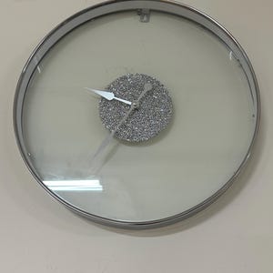 May include: A round, silver-framed wall clock with a clear glass face. The clock face is covered in silver glitter and has silver hands.