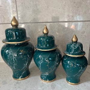 Peut inclure: Trois vases décoratifs en céramique vert foncé avec couvercles. Ils présentent des accents dorés et un motif blanc marbré. Les couvercles sont surmontés d'un bouton doré. Les vases varient en taille, le plus grand mesurant environ 30 cm de haut.