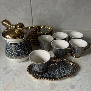 Könnte beinhalten: Ein Keramik-Kaffeeservice mit einem goldfarbenen froschförmigen Deckel auf der Kaffeekanne. Das Set umfasst kleine Tassen, Untertassen und ein Serviertablett, alle mit einem blau-goldenen Wirbelmuster.