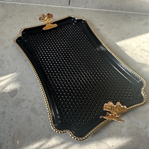 Può includere: Vassoio rettangolare nero con superficie strutturata e finiture dorate. Il vassoio ha un bordo smerlato e manici dorati a forma di foglia. Il vassoio misura circa 35 cm di lunghezza e 23 cm di larghezza.