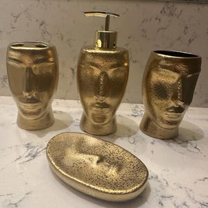 May include: A gold bathroom set with a face design. The set includes a soap dispenser, toothbrush holder, tumbler, and soap dish. The items have a textured surface and a metallic finish, adding a touch of luxury to the bathroom decor.