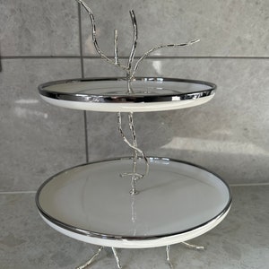 May include: Two-tiered white ceramic serving stand with a silver metal base and branches. The stand has a silver rim around each tier.