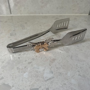 Peut inclure: Pinces de service en acier inoxydable avec un papillon et des feuilles dorées. Les pinces ont des extrémités fendues et sont conçues pour servir des aliments. Le design papillon et feuilles ajoute une touche décorative.