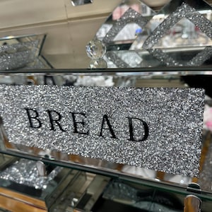 Puede incluir: Una panera de espejo plateado con un acabado brillante y brillante. La panera tiene un frente de vidrio transparente con la palabra "BREAD" impresa en negro.