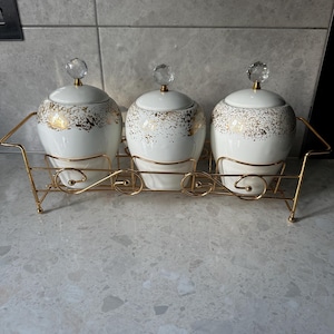 Puede incluir: Un conjunto de tres tarros de cerámica blanca con detalles dorados y tapas con cristales, exhibidos en un estante de metal dorado. Los tarros tienen forma redondeada y son adecuados para la cocina o el comedor.