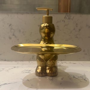 May include: A gold-colored soap dispenser with a built-in tray. The dispenser has a textured, hammered appearance and a pump top. The tray is round and sits below the dispenser. The design is modern and decorative.