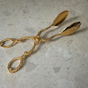 Peut inclure: Pinces de service dorées avec des détails ornés. Les pinces présentent un motif floral décoratif sur les poignées et les extrémités de service. Les pinces sont en métal brillant et réfléchissant, conçues pour servir des aliments.