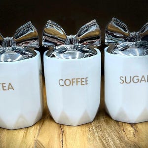 May include: Three white ceramic canisters with silver bow lids. The canisters are labeled "Tea", "Coffee", and "Sugar".