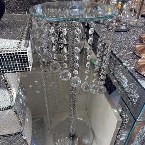 Op de afbeelding: Een zilveren spiegelsokkel met een glazen bovenblad en kristallen strengen die eronder hangen. De sokkel is versierd met strass.
