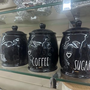 May include: Three black ceramic canisters with heart-shaped lid knobs. The canisters are labeled "TEA", "COFFEE", and "SUGAR" in white script. The canisters are on a glass shelf.