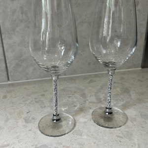 May include: Two clear wine glasses with crystal-studded stems. The glasses have a classic, rounded bowl shape and sit on a clear, circular base. The stems are decorated with small, sparkling crystals. The background is a light grey.