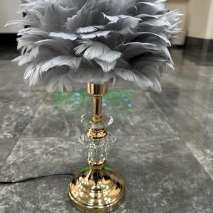 Op de afbeelding: Een tafellamp met een pluizige, lichtgrijze veren lampenkap. De lampvoet is gemaakt van gepolijst goudkleurig metaal met een centraal, gefacetteerd glazen decoratief element. Een zwart netsnoer komt uit de ronde, gouden voet.