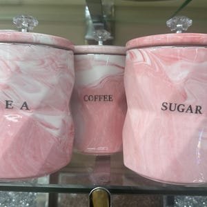 Może przedstawiać: Trzy różowo-białe pojemniki z wzorem marmuru i pokrywkami z kryształowymi gałkami. Pojemniki są oznaczone napisami "TEA", "COFFEE" i "SUGAR" czarnym tekstem. Pojemniki są wystawione na szklanej półce.