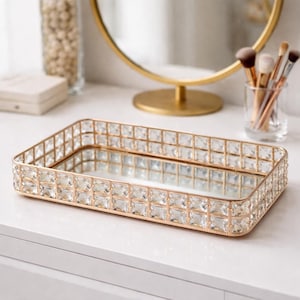Könnte beinhalten: Ein rechteckiges, goldfarbenes Tablett mit verspiegeltem Boden und einer Umrandung aus klaren, quadratisch geschliffenen Kristallen. Das Tablett steht auf einer weißen Oberfläche, mit einem goldgerahmten Spiegel und Make-up-Pinseln im Hintergrund. Ein dekorativer Artikel für die Kommode.
