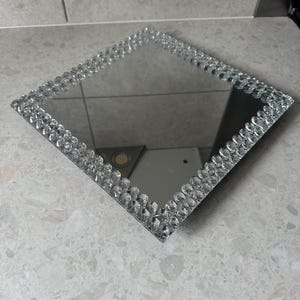 Könnte beinhalten: Ein quadratisches Spiegeltablett mit einem klaren Kristallrand. Das Tablett ist perfekt zum Präsentieren kleiner Gegenstände oder zum Servieren von Snacks.