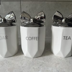 Könnte beinhalten: Drei weiße Keramikdosen mit silbernen Schleifen-Deckeln. Die Dosen sind mit "Zucker", "Kaffee" und "Tee" beschriftet.