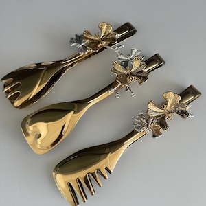 May include: Three gold-coloured serving utensils with a floral design. Each utensil has a different end: a fork, a spoon, and a comb-like shape. The floral design is gold and silver. The utensils are on a white surface.
