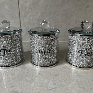 Op de afbeelding: Drie zilveren glitterpotten met doorzichtige deksels en kristalachtige knoppen. De potten zijn gelabeld met "Koffie", "Suiker" en "Thee".
