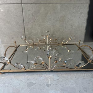 May include: A rectangular gold-toned mirrored tray with a decorative crystal and gold floral design. The tray has a mirrored surface and is accented with clear, teardrop-shaped crystals. The design is symmetrical and elegant.