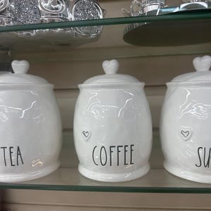May include: Three white ceramic canisters with heart-shaped lid knobs. The canisters are labeled "TEA", "COFFEE", and "SUGAR" in black text. Each canister has a small heart design. The canisters are on a glass shelf.