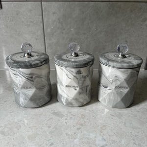 Pode incluir: Três potes de cerâmica cinza com padrões de mármore e botões de cristal transparente. Os potes são etiquetados com "Chá", "Açúcar" e "Café".