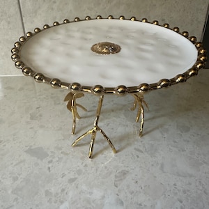 May include: White ceramic cake stand with a gold rim and a gold decorative centrepiece. The stand has three gold metal legs that resemble tree branches.