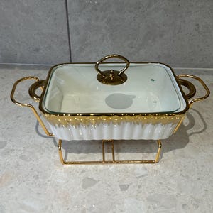 May include: A white and gold rectangular casserole dish with a clear glass lid and gold handle. The dish has a gold rim and sits in a gold metal stand. The dish is designed for serving food.