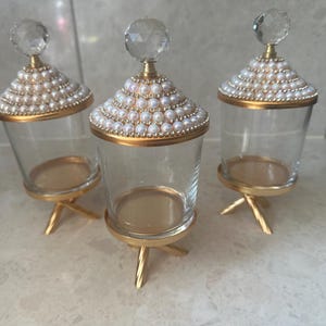 Könnte beinhalten: Drei transparente Glasbehälter mit goldfarbenen Böden und Deckeln. Die Deckel sind mit Kunstperlen verziert und mit einem kristallartigen Knauf versehen. Die Gläser sind für die dekorative Aufbewahrung bestimmt.