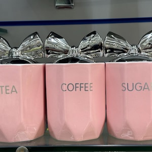 Könnte beinhalten: Drei rosa Keramikdosen mit silbernen Schleifenakzenten. Die Dosen sind mit "TEA", "COFFEE" und "SUGAR" beschriftet.