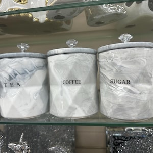 May include: Three white ceramic canisters with marbleised grey accents and silver lids. The canisters are labelled "Tea", "Coffee", and "Sugar".