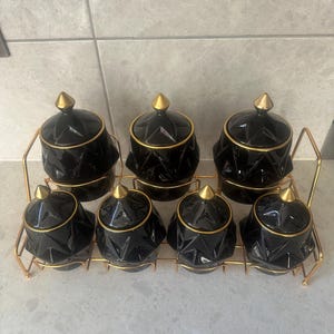Pode incluir: Um conjunto de oito potes de especiarias de cerâmica preta com detalhes dourados, dispostos em uma prateleira de metal dourado. Cada pote tem um design geométrico e uma tampa dourada. Os potes são projetados para uso na cozinha.