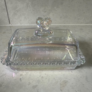 Könnte beinhalten: Eine klare Glasbutterdose mit einem herzförmigen Deckel. Die Schale hat einen perlenbesetzten Rand und eine irisierende Oberfläche.