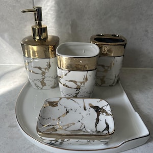Può includere: Un set da bagno di quattro pezzi con un design a marmo bianco e dettagli dorati. Il set include un dosatore di sapone, un portaspazzolino, un bicchiere e un portasapone, il tutto disposto su un vassoio bianco.