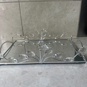 May include: A rectangular mirrored tray with a silver metal frame and decorative crystal accents. The tray features curved handles and a floral-like design with clear crystals. The crystals are teardrop-shaped and add a touch of elegance.