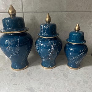 May include: Three decorative ceramic jars in varying sizes. The jars are a deep teal color with a marble-like white and gold veining pattern. Each jar has a matching lid with a gold finial and gold trim.