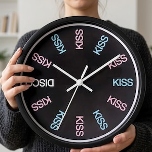 May include: Black round wall clock with white hands and the word "DISCO" in white. The word "KISS" is printed in pink and light blue around the clock face. The clock is held by a person wearing a gray sweater.